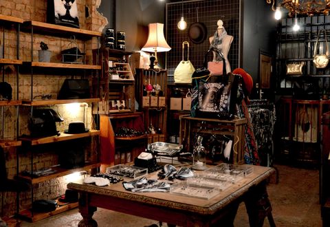 A dark vintage accessory store lit by lamps and bare bulbs, with bags, jewelry, sunglasses and a bowler hat on the wooden shelves and carved table.
