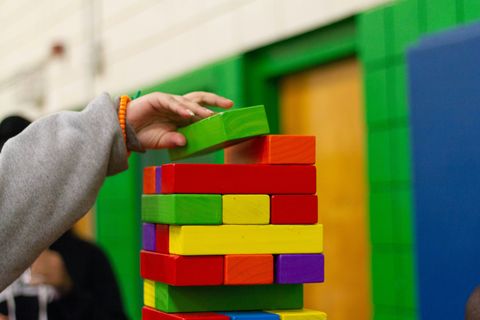 A hand with an orange bracelet extends from a baggy gray sweatshirt and places a large green block on top of a colorful and precarious stack of Jenga-like blocks.
